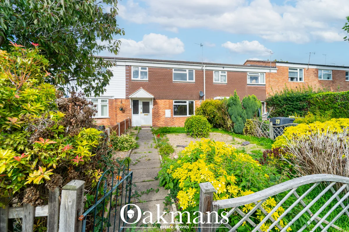 Front garden with yellow flowering plants, green shrubs, wooden fence, pathway to brick terraced house with white entran
