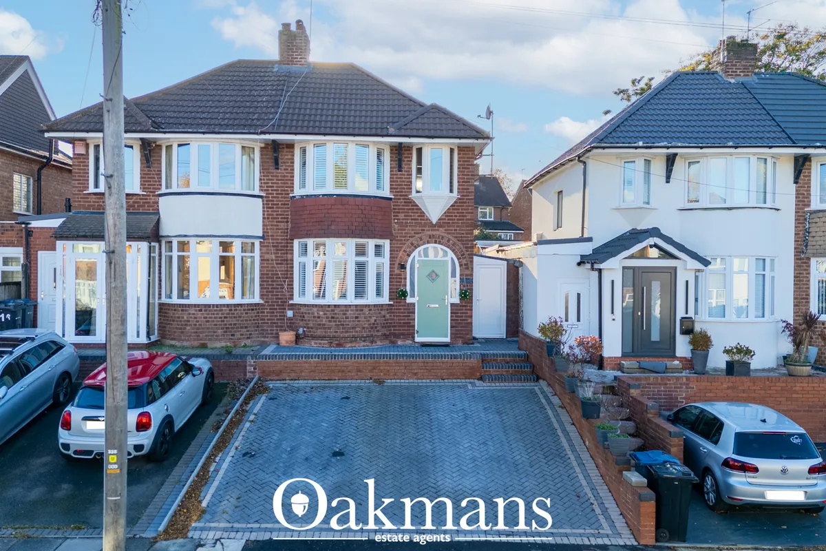 Front view of red-brick semi-detached house with bay windows, dark roof tiles, driveway with parked cars, and neighbouri