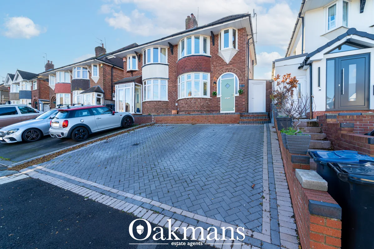 Red brick semi-detached house with bay windows, driveway with two parked cars, paved forecourt, and neighbouring propert