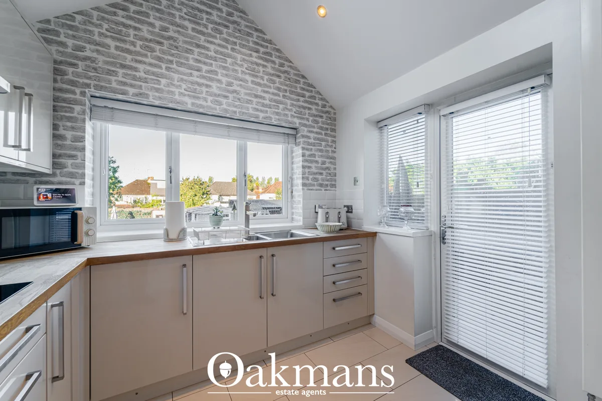 Modern fitted kitchen with beige cabinetry, wood worktops, dual sink, brick feature wall, grey blinds and large windows 