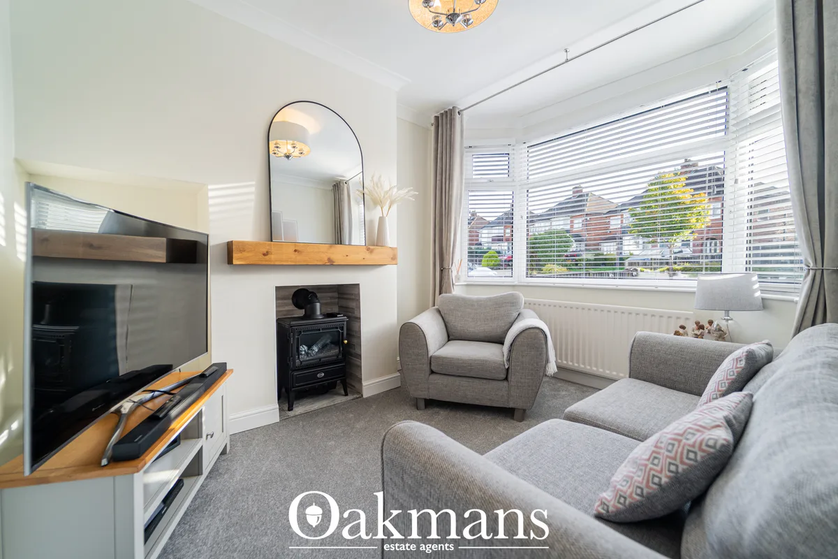 Bright living room with grey upholstered sofa and armchair, wood-burning stove, TV unit, large bay windows with shutters