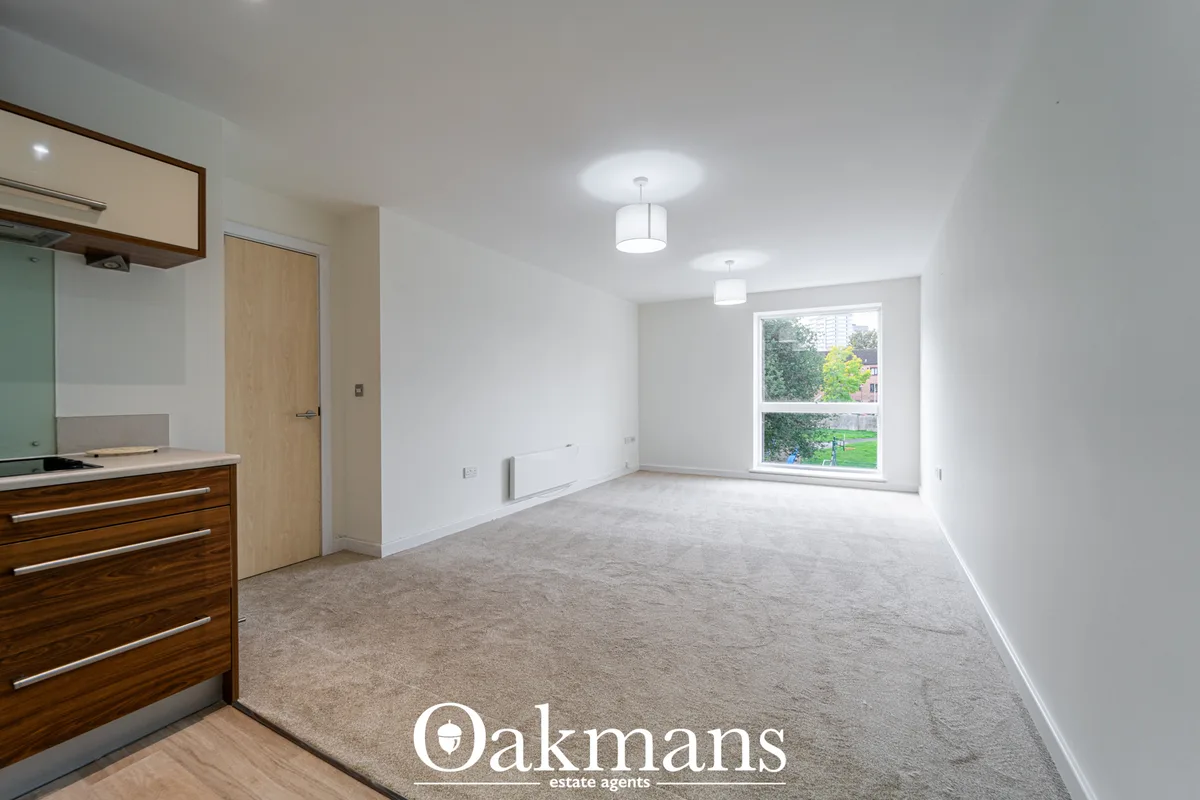 Bright open-plan living space with fitted kitchen to left, large window overlooking garden, beige carpet, pendant ceilin