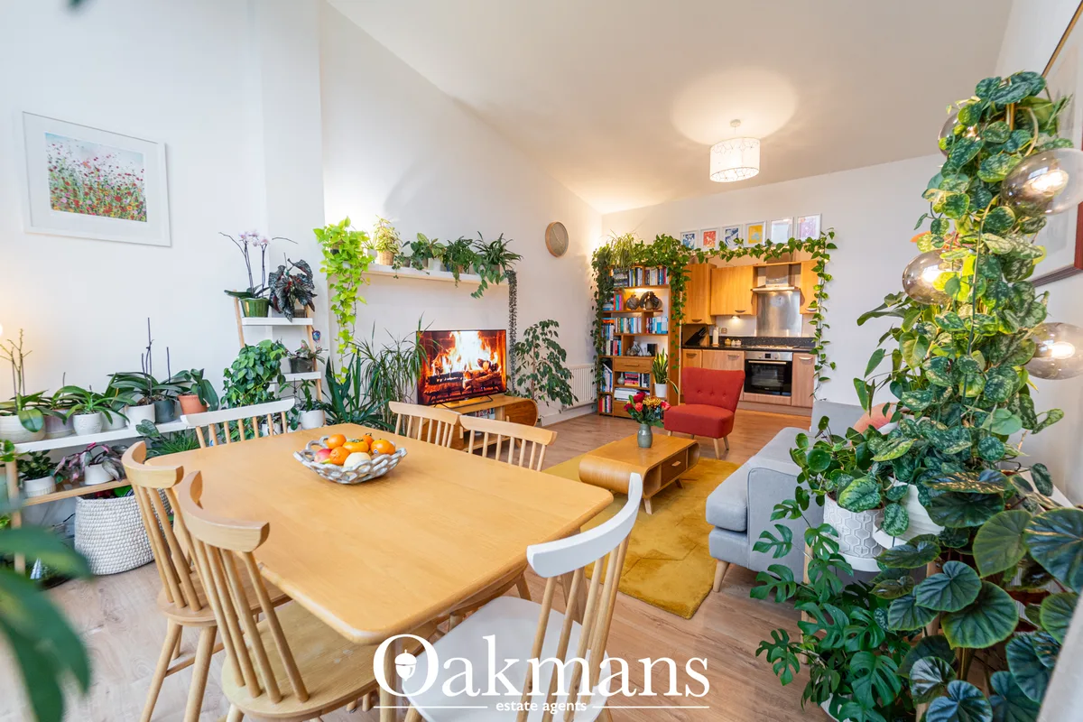 Open-plan living space with wooden dining table, light wood chairs, potted plants throughout, fireplace with logs, red a
