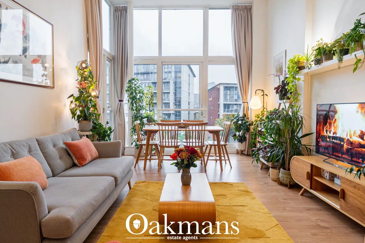 Bright open-plan living room with grey sofa, wooden dining table, large windows overlooking city, potted plants and wood
