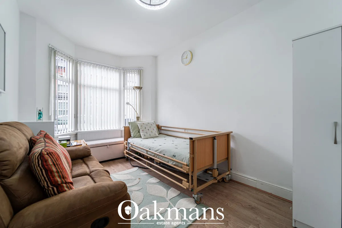Bright bedroom with wooden hospital bed, brown leather armchair, vertical blinds, wall-mounted clock and wooden flooring