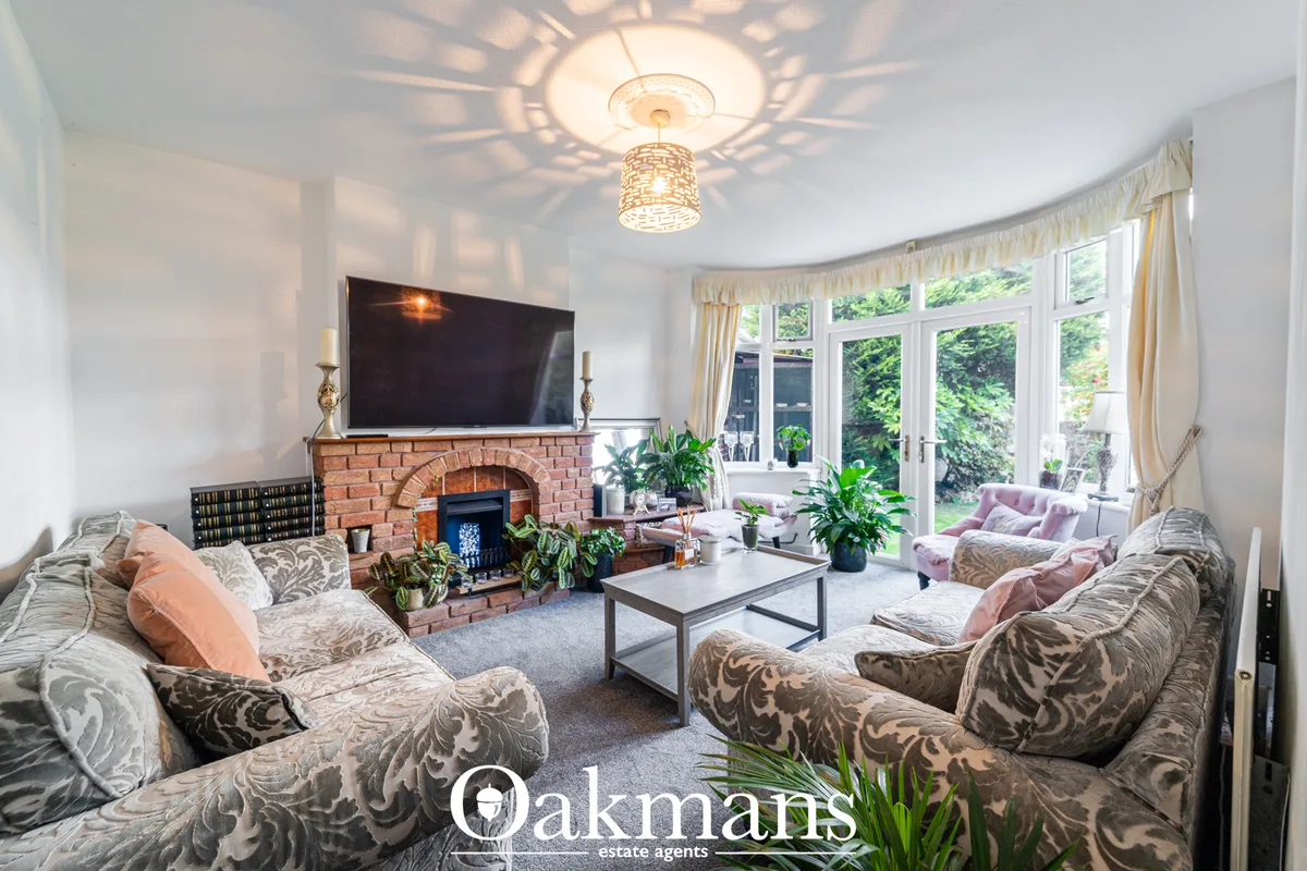 Spacious living room with patterned sofas, brick fireplace with TV, ornate ceiling light, French doors overlooking garde
