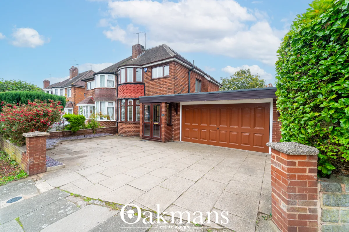 Red brick semi-detached house with brown garage door, paved driveway, manicured gardens with hedges and flowering shrubs