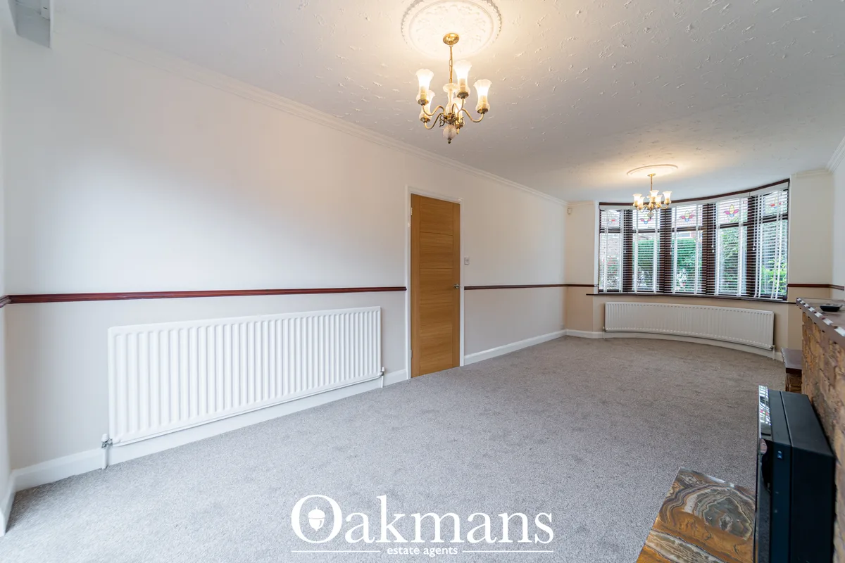 Spacious living room with cream walls, grey carpet, white radiator, wooden door, large bay window with shutters and bras