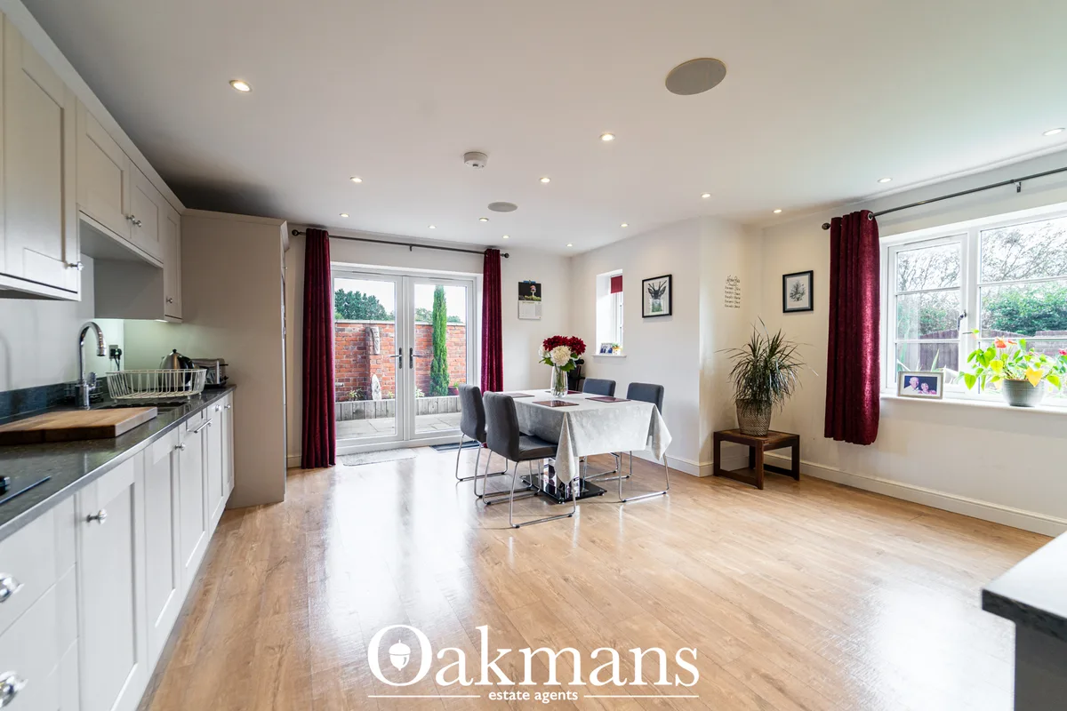 Open-plan dining and kitchen area with white fitted units, dining table with grey chairs, burgundy curtains, wooden floo