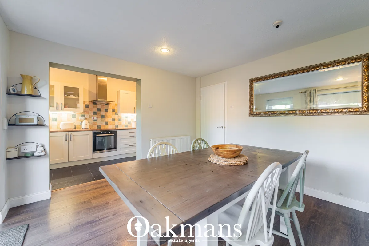 Dining room with wooden table, white chairs, decorative bowl and mirror. Open-plan to fitted kitchen with cream units an