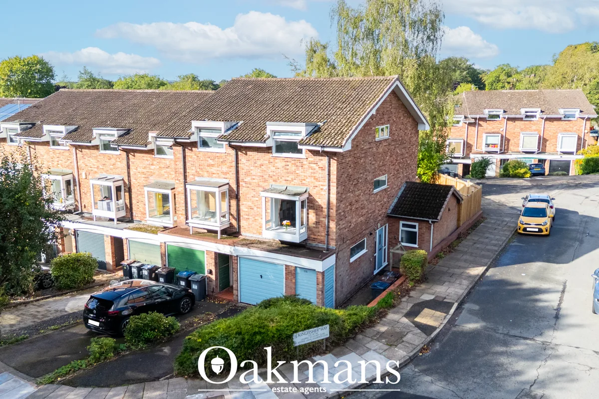 Aerial view of brick terraced houses with tiled roofs, driveways, garages, parked cars, surrounding trees and blue sky