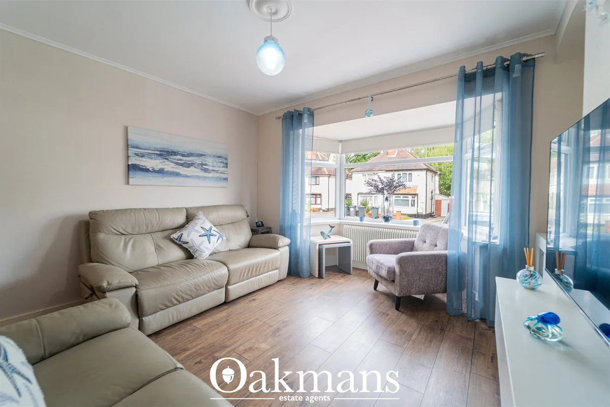 Bright living room with beige leather sofa, grey armchair, blue sheer curtains, wooden flooring, and large windows overl