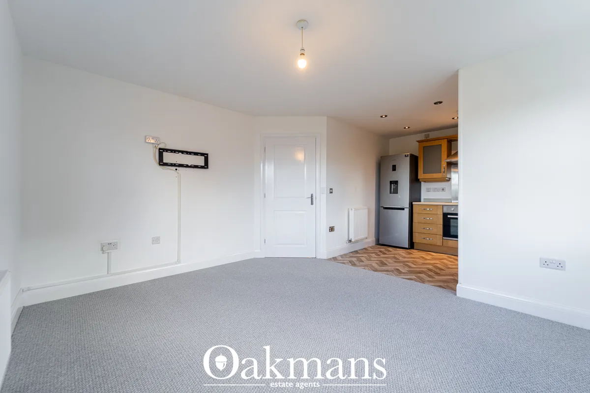 Spacious open-plan living room with grey carpet, white walls, ceiling pendant light, and kitchen visible through archway