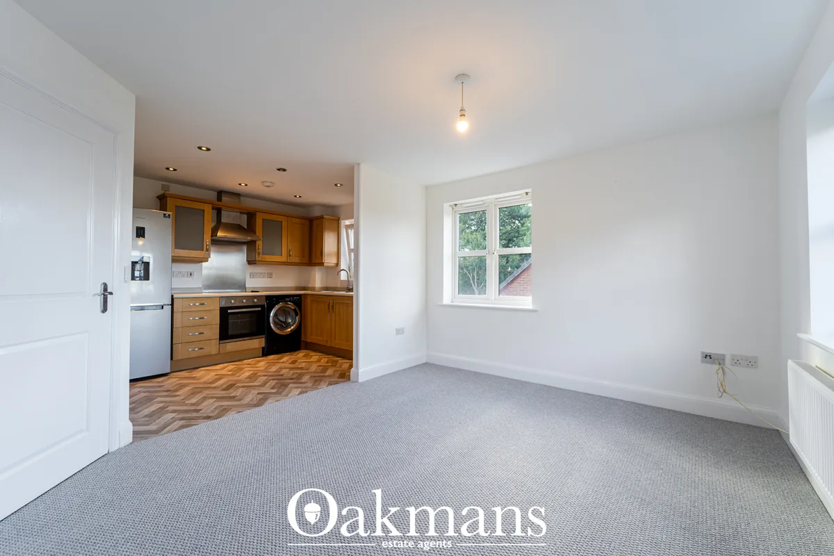 Open-plan living and kitchen area with grey carpet, white walls, wooden kitchen units, window with garden view
