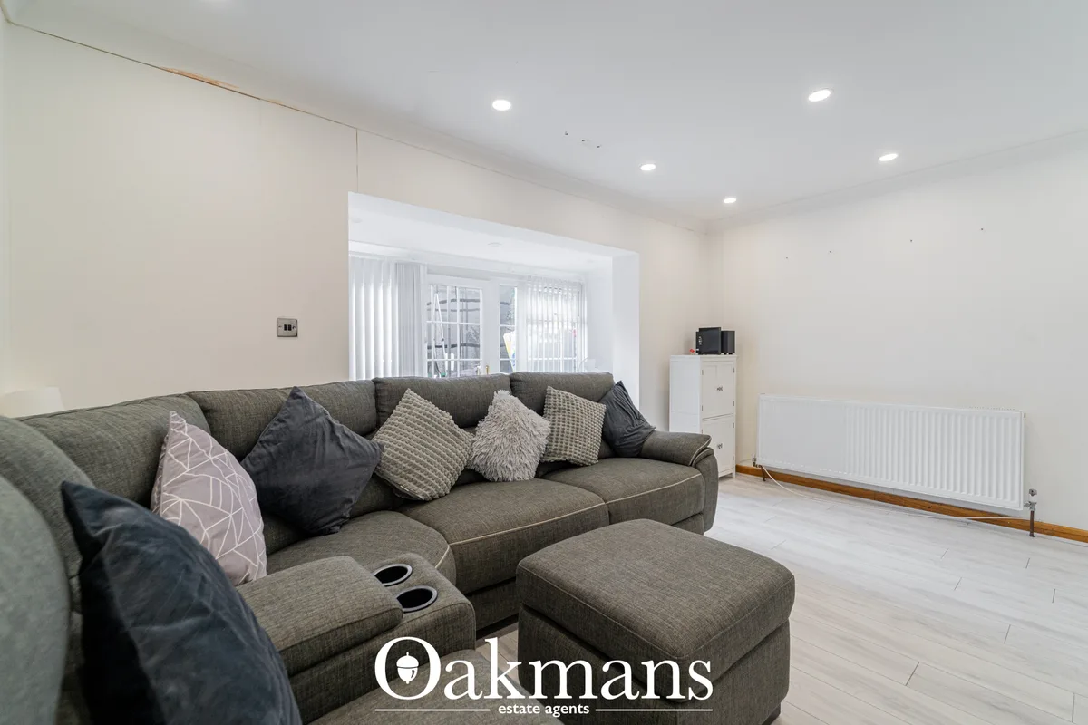 Spacious living room with grey corner sofa, scatter cushions, footstool, white radiator, vertical blinds on patio doors 