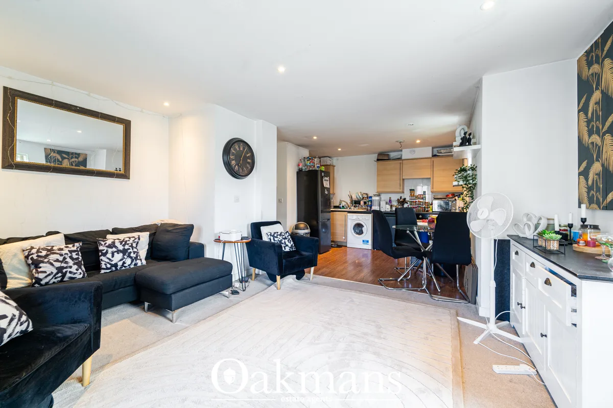 Open-plan living room with black sofa, armchairs, patterned cushions, wooden flooring, wall mirror, and kitchen area vis