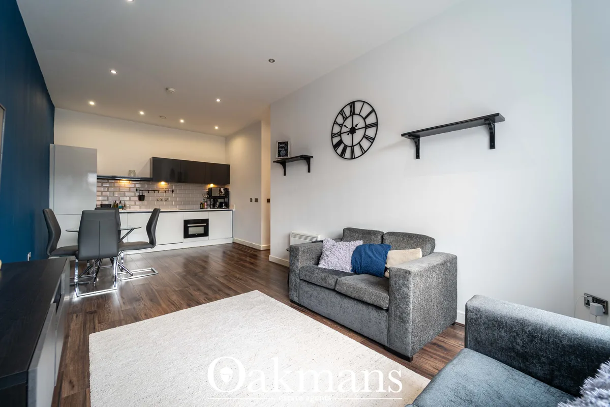 Open-plan living room with grey sofas, white rug, wooden flooring, wall-mounted clock and shelving, with kitchen area vi