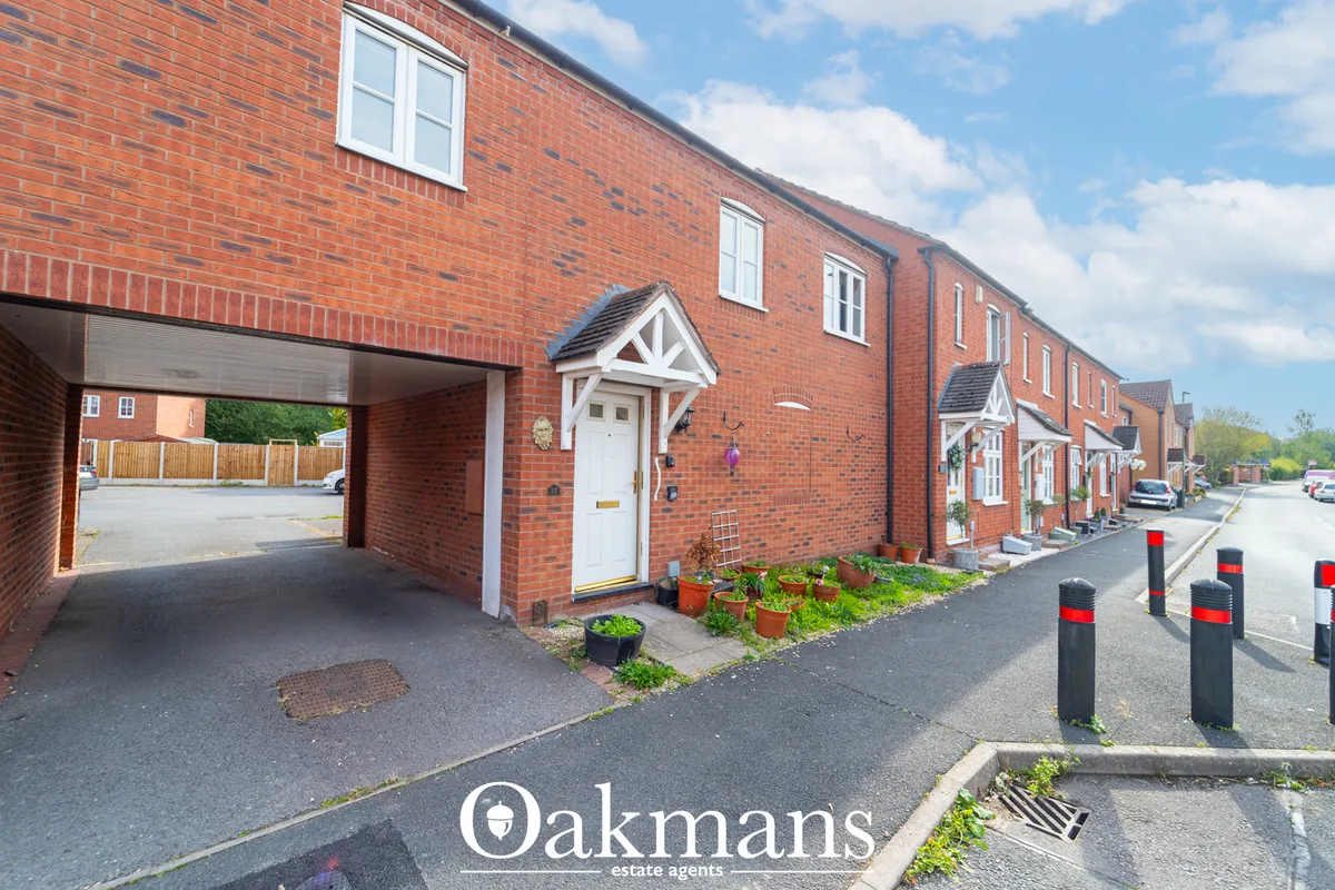 Red brick terraced house with white entrance door, garage, multiple windows, potted plants, and street with bollards