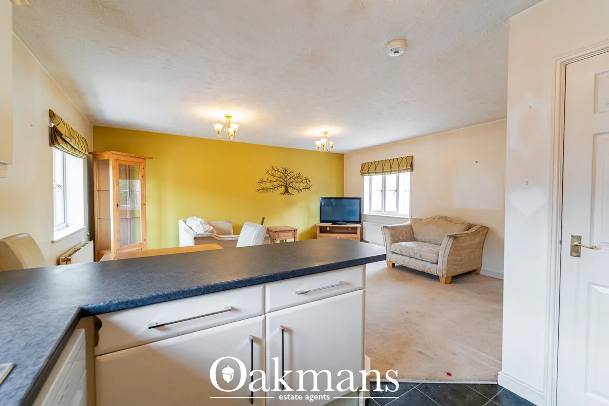 Open-plan living room with yellow feature wall, beige armchair, TV, kitchen with dark worktops, and multiple windows wit