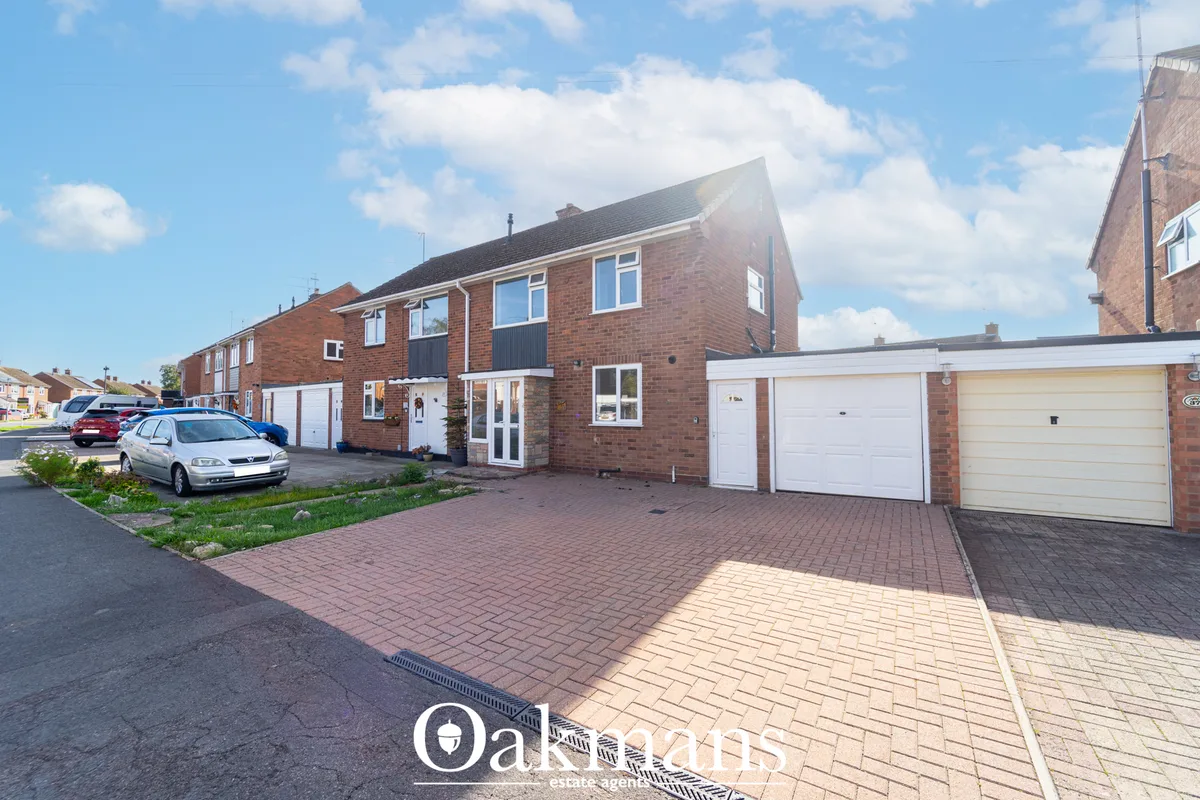 Red brick semi-detached house with driveway, parked cars, brick paved forecourt and white garage doors