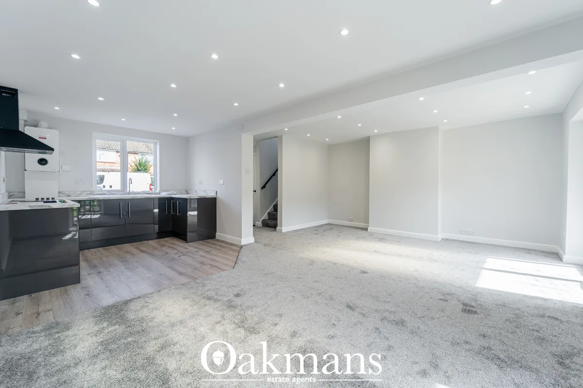Open-plan living room with pale grey carpet, white walls, recessed ceiling lights, and black kitchen units visible in ba