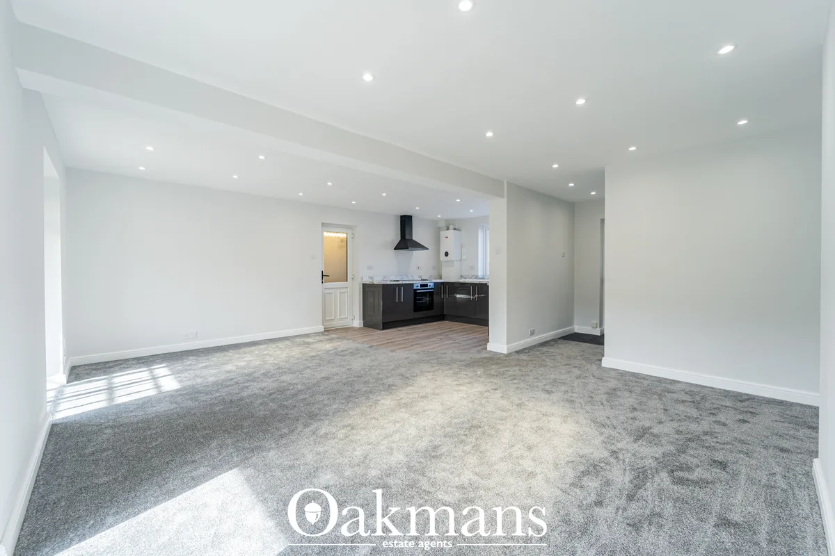 Spacious open-plan living area with grey carpet, white walls, recessed ceiling lights, and fitted black kitchen unit in 