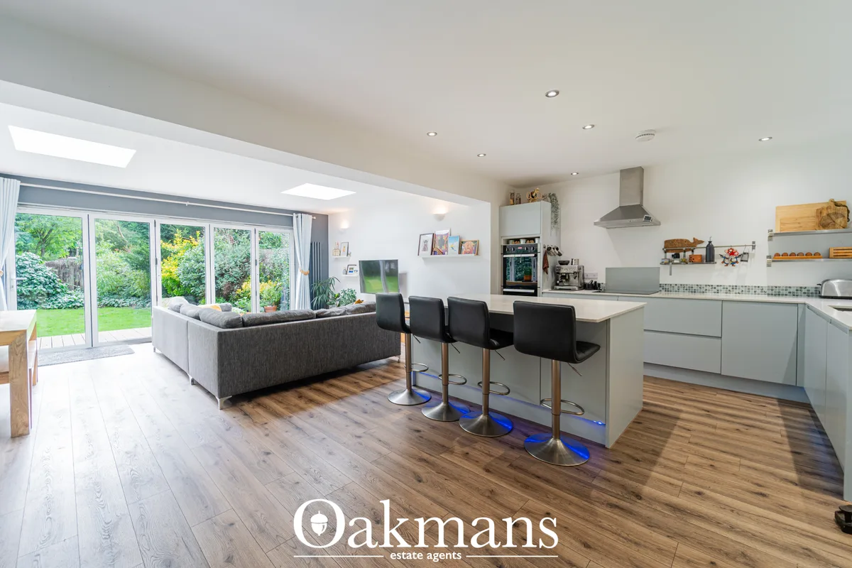 Open-plan living room with grey sofa, bi-fold doors opening to garden, skylights, and integrated kitchen with white unit