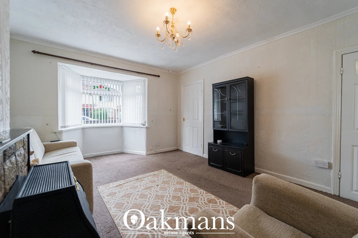 Spacious living room with beige walls, bay window with vertical blinds, cream carpet, patterned rug, beige upholstered s
