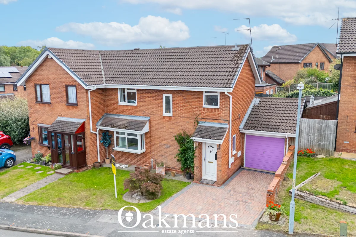 Red brick detached house with purple garage door, driveway, landscaped front garden with shrubs and lawn, parked cars vi