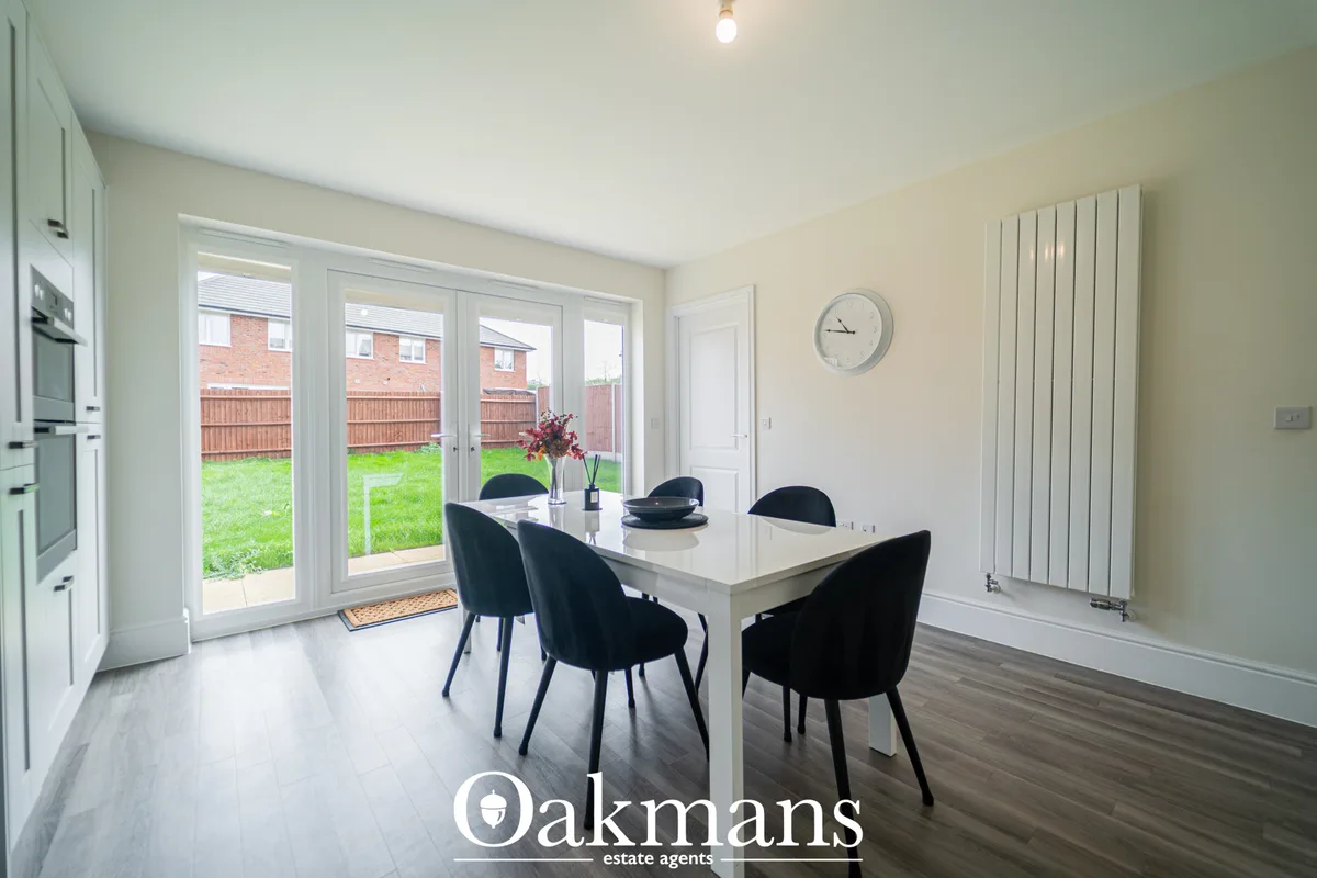 Modern dining room with white table, black chairs, French doors opening to garden, white kitchen units visible left, ver