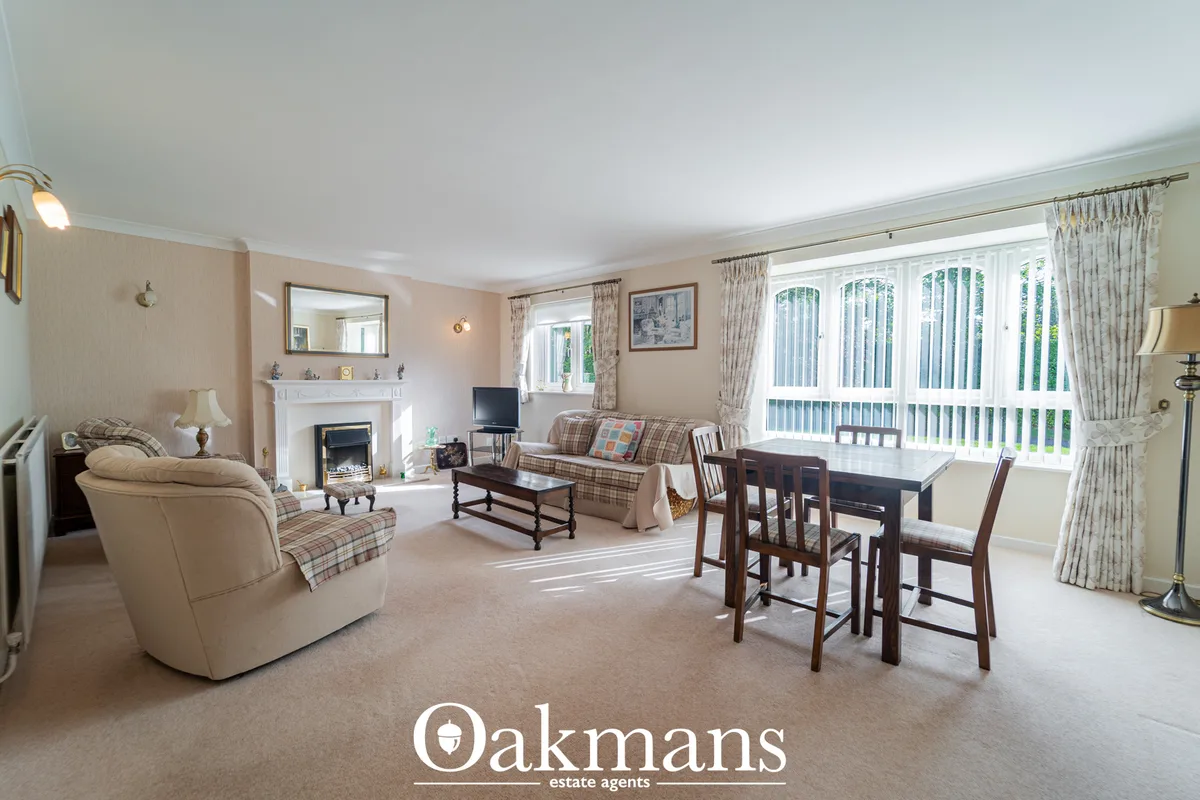 Spacious living room with beige walls, fireplace with stove, cream armchair, patterned sofa, dining table with chairs, l
