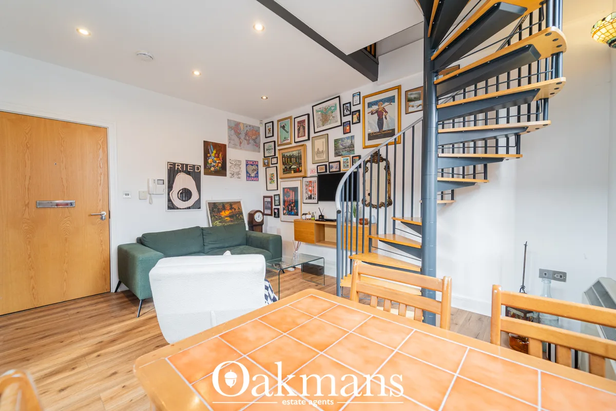 Open-plan living space with green sofa, dining table, wooden staircase with metal railings, gallery wall art, and mezzan
