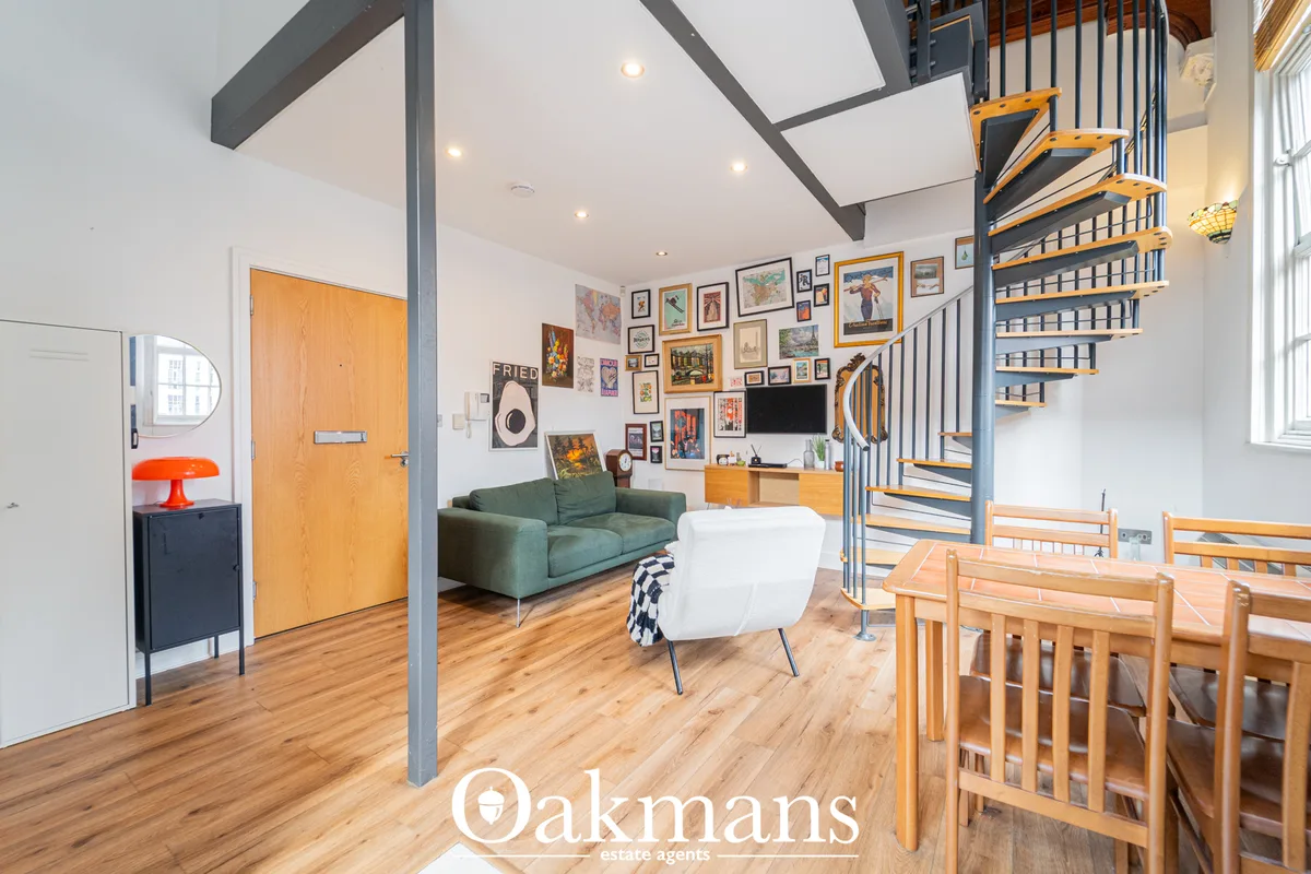 Open-plan living space with green sofa, white armchair, wooden dining table, exposed grey beams, wood flooring, gallery 