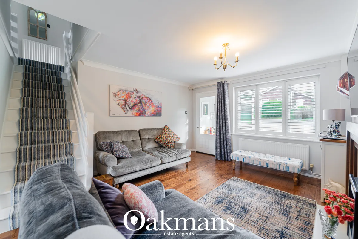 Bright living room with grey sofas, wooden flooring, wall art, large windows with shutters, hallway with stairs visible 