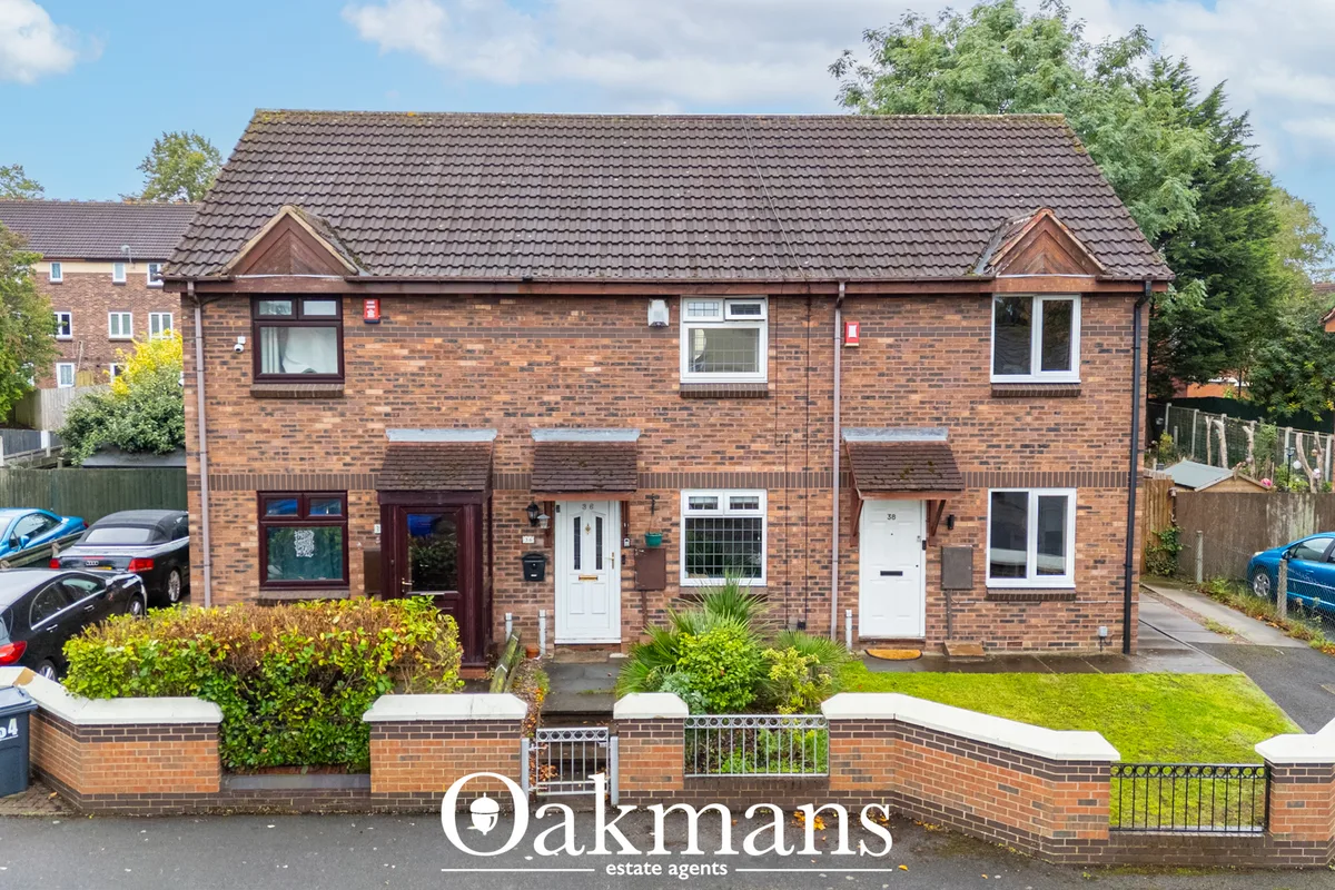 Brick-built semi-detached house with dark tile roof, front gardens with hedging, driveway and parked cars