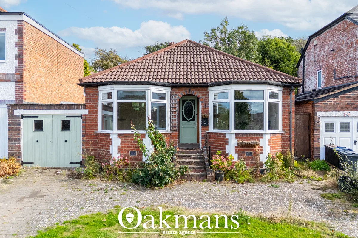 Red brick bungalow with hipped tile roof, white bay windows, green door, gravel driveway and adjoining garage