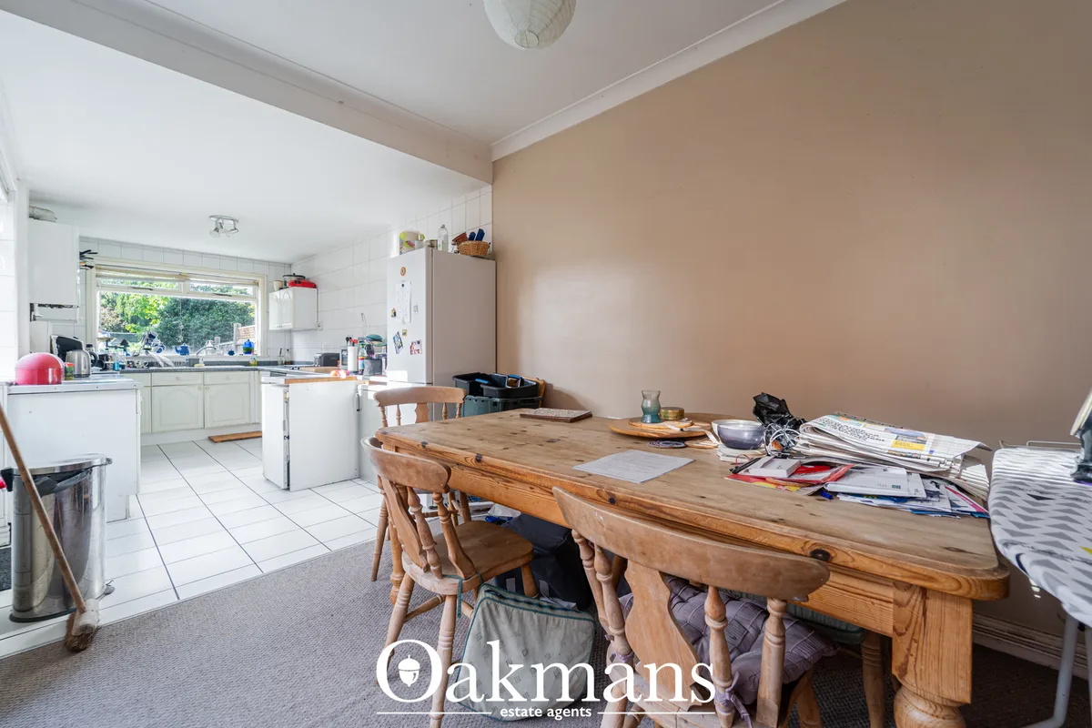 Open-plan dining and kitchen area with wooden dining table, chairs, beige wall, white ceiling and natural light from lar