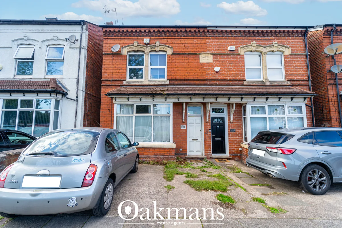 Red brick two-storey semi-detached house with cream stone window surrounds, black door, parked cars in front