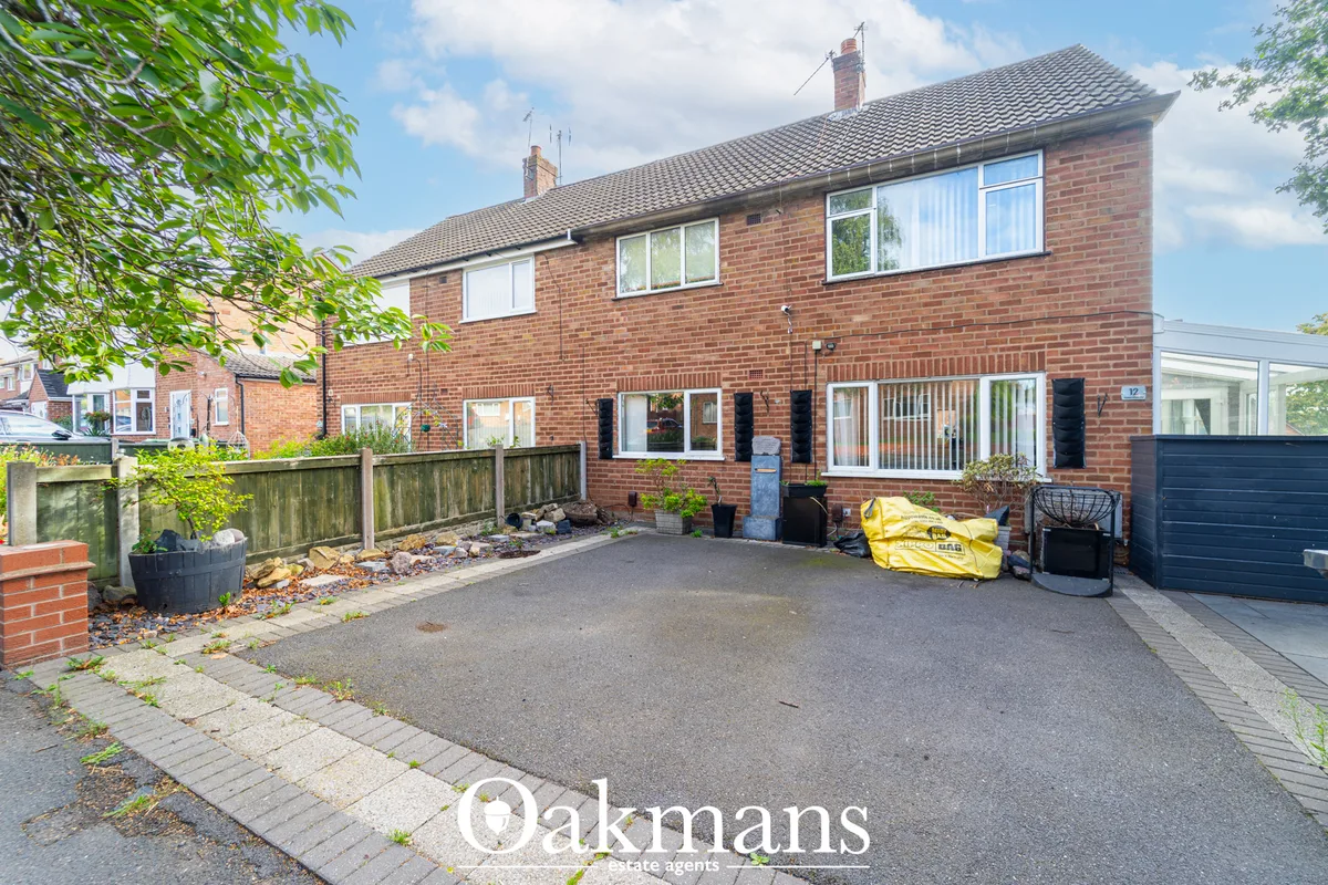 Red brick semi-detached house with driveway, garage, multiple windows, and mature trees framing the front façade