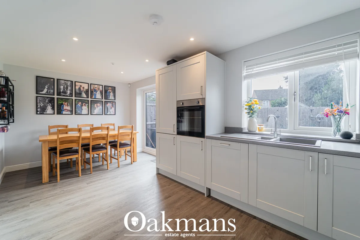 Modern open-plan kitchen with cream fitted units, dark worktops, integrated oven, and stainless steel sink. Wooden dinin