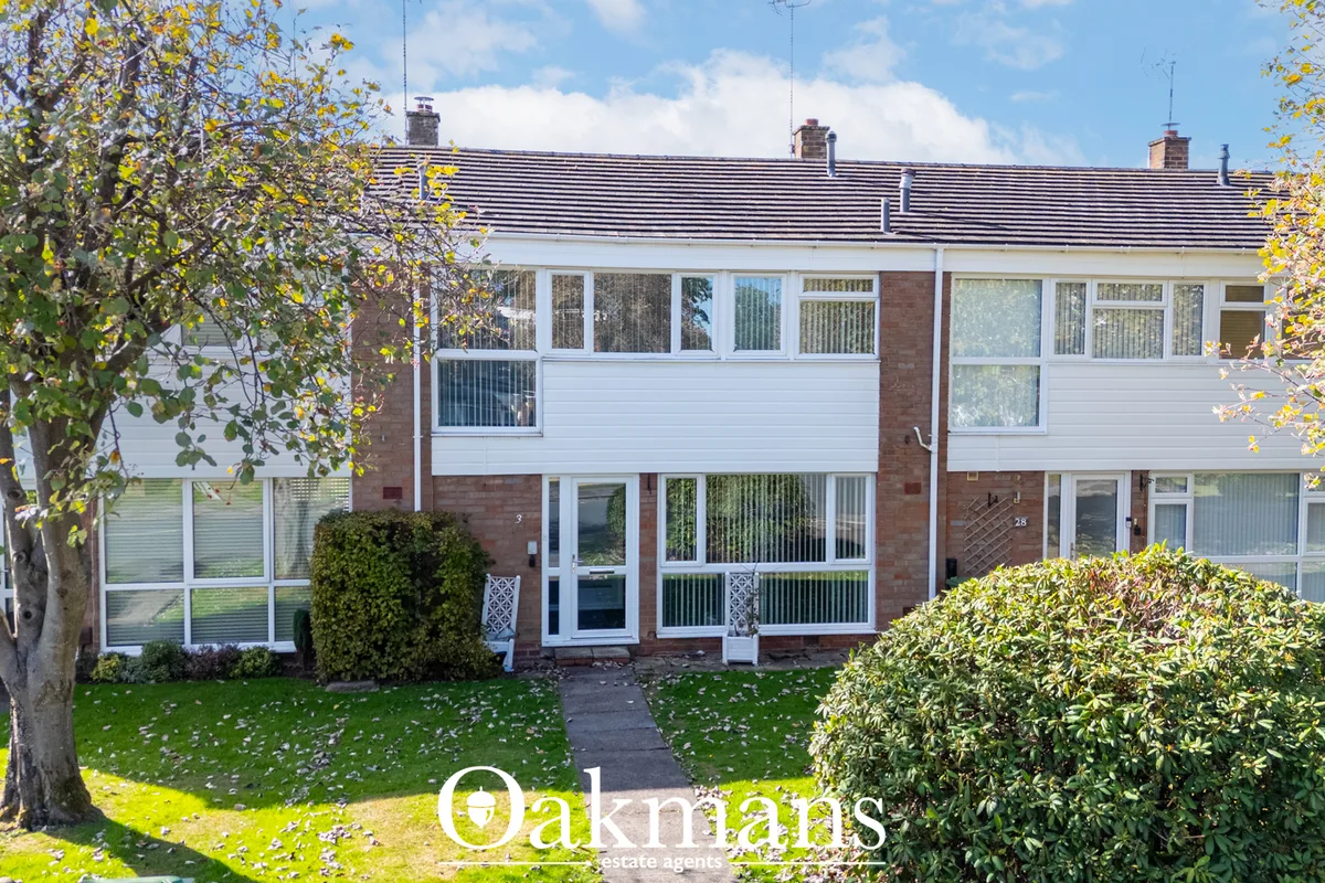 Two-storey brick terraced house with white UPVC cladding, multiple windows, pitched roof with chimneys, front garden wit