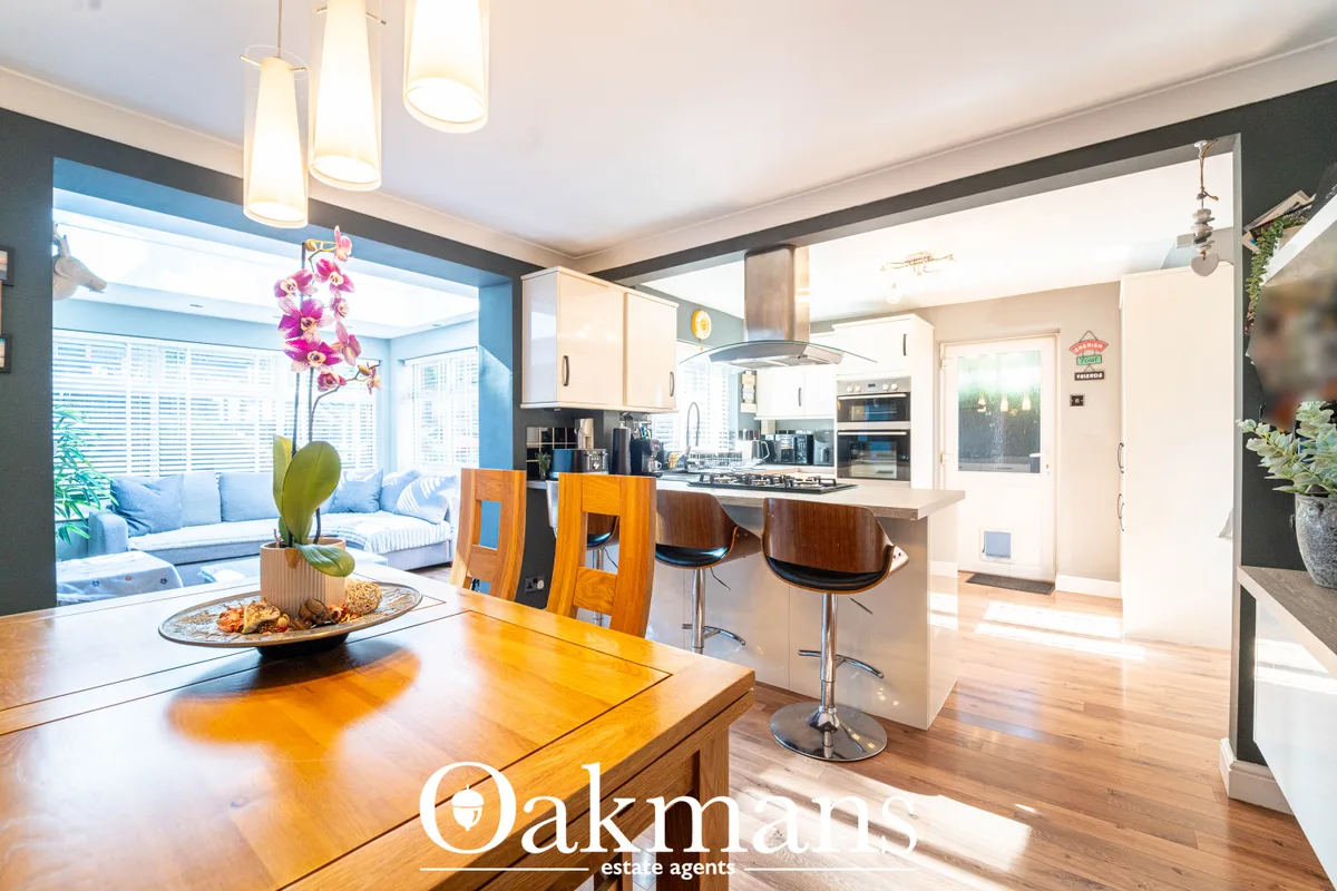 Modern open-plan kitchen with white units, wooden dining table, breakfast bar with stools, and view into conservatory wi