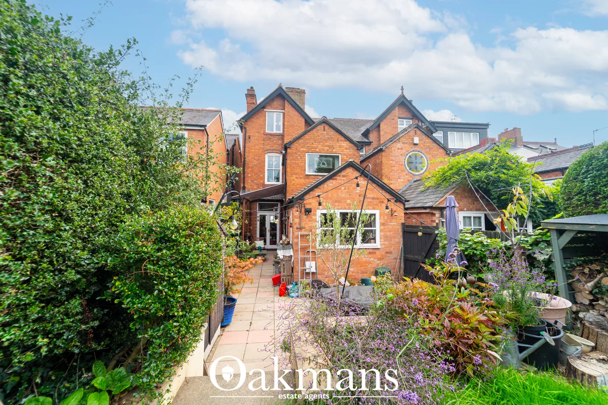 Rear garden with paved patio, mature hedging, flowering plants, brick Victorian property facade visible