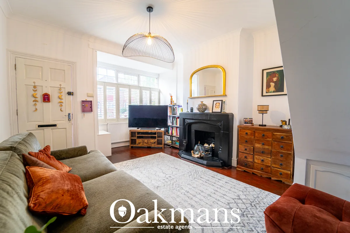 Bright living room with cream walls, wood flooring, grey sofa with orange cushions, black stove fireplace, wooden chest 
