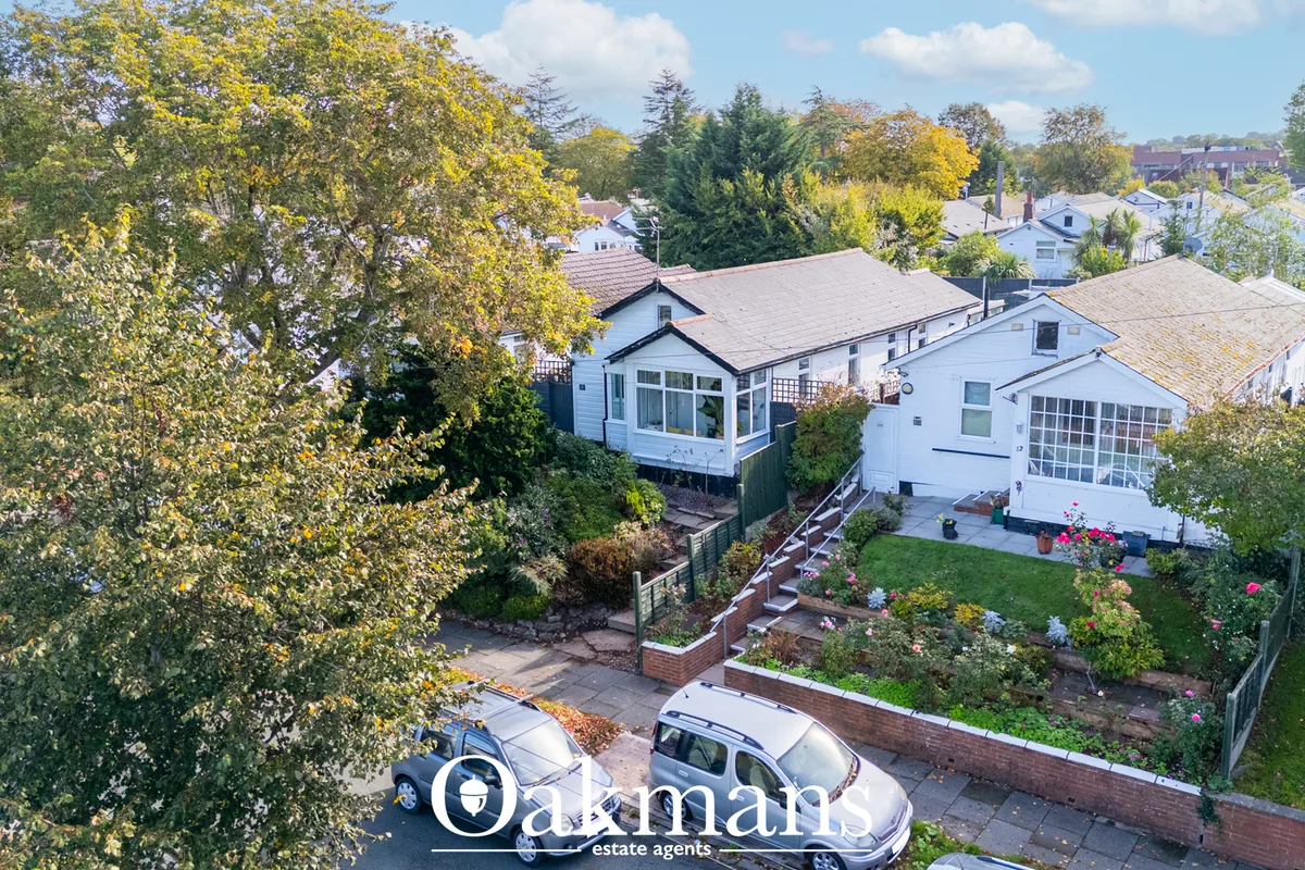 Aerial view of white bungalow with gray roof, front garden with flowering borders, driveway with two cars, mature trees 