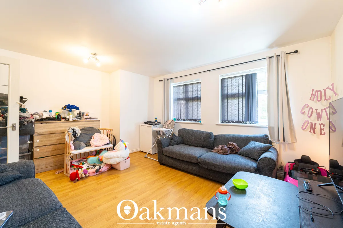Spacious living room with grey sofas, wooden flooring, multiple windows with blinds and curtains, dresser, cot, toys and