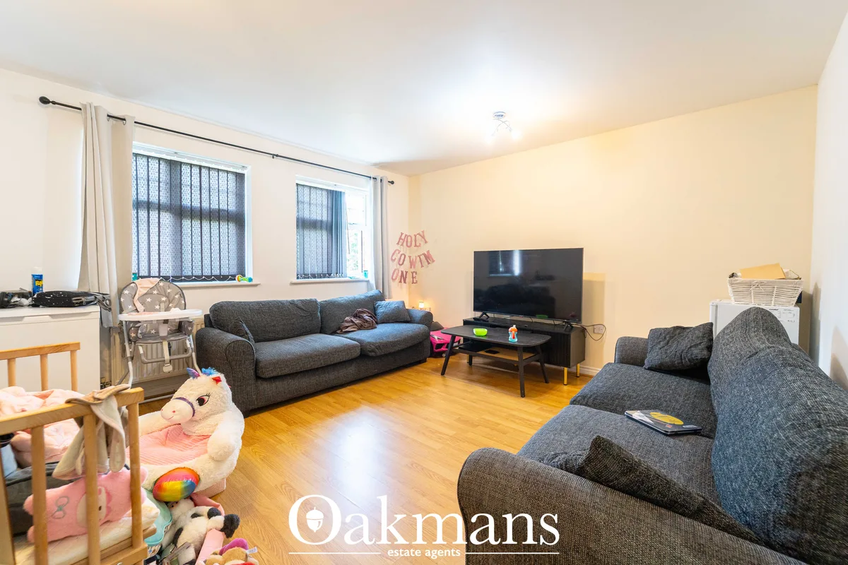 Spacious living room with two grey sofas, flat-screen TV on stand, wooden flooring, large windows with blinds, cream wal