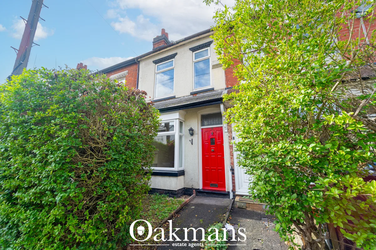 Red-brick terraced house with cream render and red front door, flanked by green hedges, clear blue sky