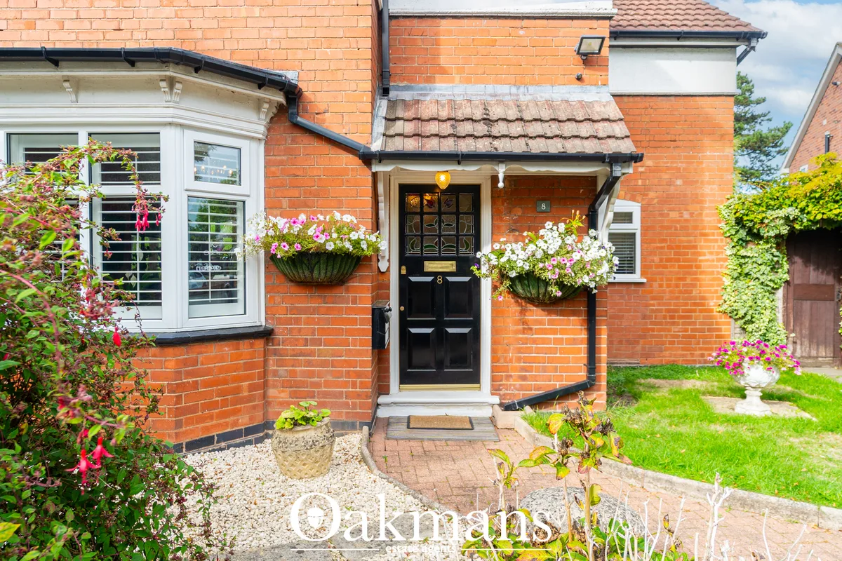 Red brick semi-detached house with black entrance door, white bay window, hanging baskets with pink and white flowers, a