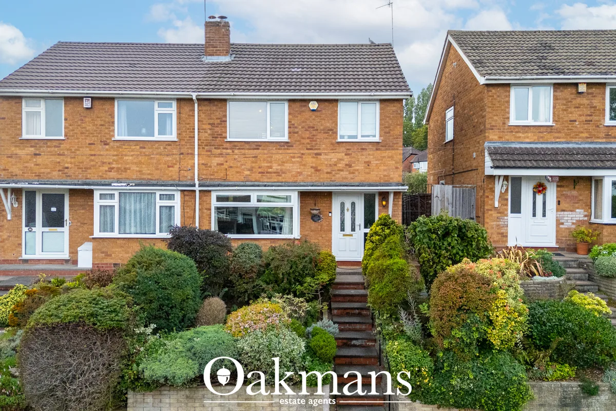 Two-storey brick semi-detached house with tiled roof, white-framed windows and doors, mature landscaped front garden wit